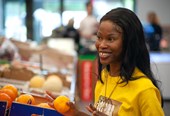 Michelle, a young woman with black hair is facing to the right and smiling broadly. She is wearing a yellow t-shirt and a gold necklace, and is holding an orange in her right hand. Behind her is a blurred background of a fruit and vegetable display.