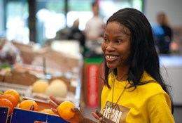 Michelle, a young woman with black hair is facing to the right and smiling broadly. She is wearing a yellow t-shirt and a gold necklace, and is holding an orange in her right hand. Behind her is a blurred background of a fruit and vegetable display.